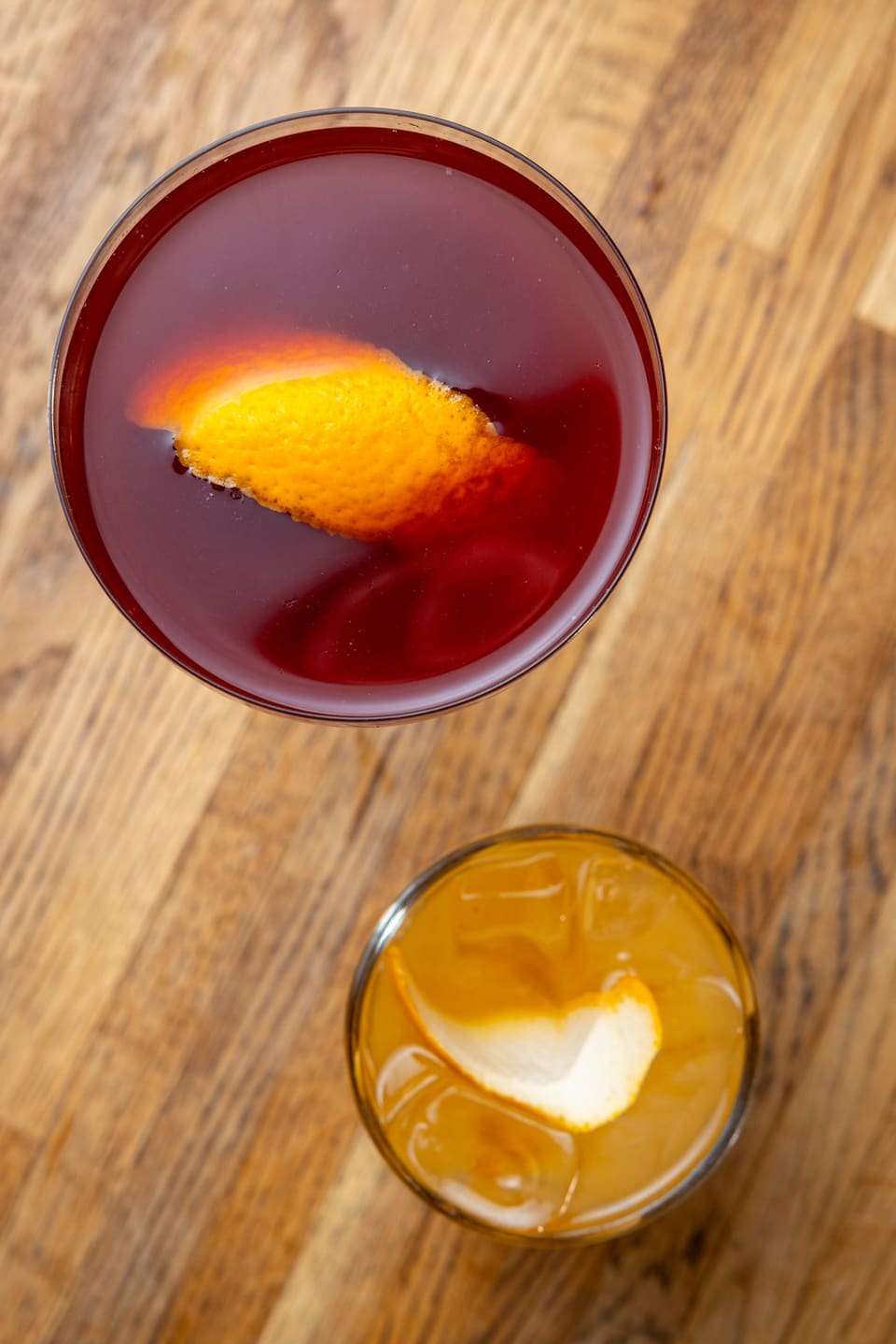A red coupe cocktail with an orange twist and an amber cocktail on the rocks